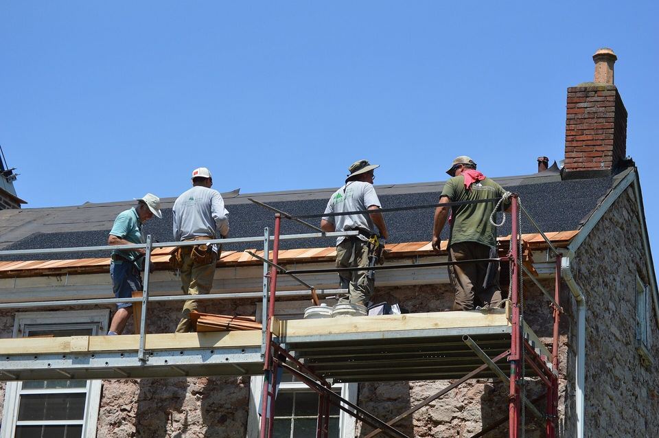 Roofers on scaffolding setting the first course of cedar shakes along a stone house eave
