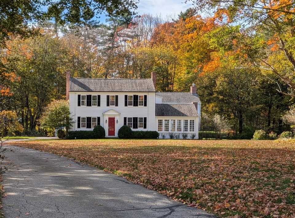 White colonial house with black shutters and a red door among bright autumn trees