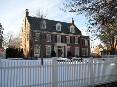 Red brick colonial house with dormers and a dark roof behind a white fence in snow