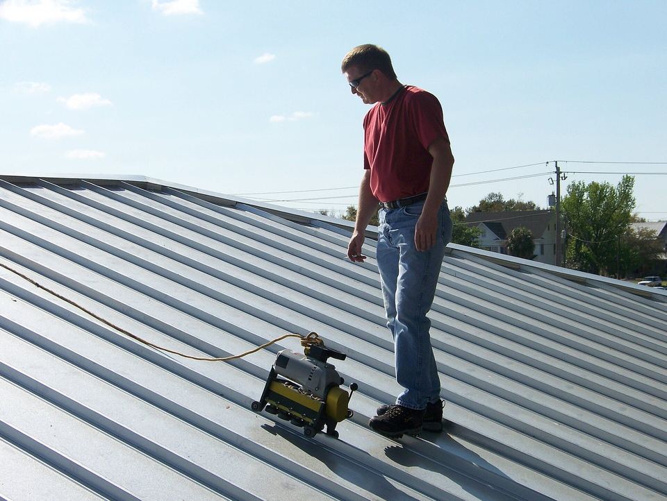 Worker on a gray standing-seam metal roof watching an electric seaming machine