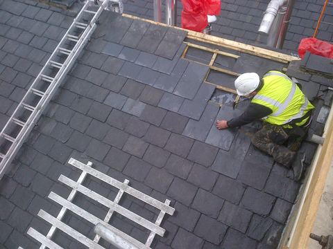 Roofer in a hard hat fitting replacement slates into a gray slate roof