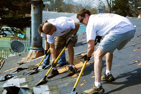 Two workers using roofing shovels to strip worn asphalt shingles off a house roof