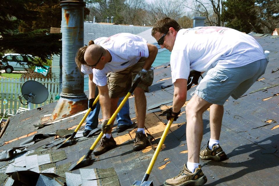 Two workers using roofing shovels to strip worn asphalt shingles off a house roof